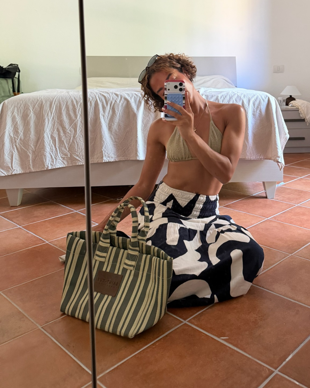 A person takes a mirror selfie in a bedroom while wearing a beige ribbed crop top and black-and-white patterned pants. A striped tote bag sits on the terracotta tiled floor beside them.
